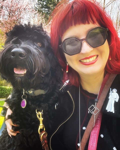 A black Portuguese water dog and a white woman woman with bright cherry red hair and lipstick and large grey-lensed sunglasses smile together with trees and grass in the background
